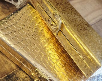 May include: Two rectangular boxes with metallic gold finishes. The bottom box features a textured crocodile skin pattern, while the top box displays an intricate embossed floral design. Both boxes are secured with a gold-toned metal latch.
