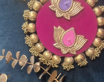 May include: A circular magenta decorative base adorned with gold-toned beads and metallic lotus-shaped candle holders. Two lotus candles in purple and pink sit on the base, while two orange lotus candles are placed nearby.