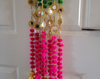 May include: Decorative garland with green and hot pink pom-poms, gold-colored mirror accents, and gold bells. The garland is hanging against a white door. The pom-poms are arranged in vertical strands.