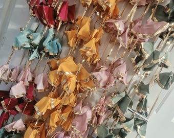 May include: A decorative hanging garland featuring multiple fabric flower-like shapes in shades of pink, yellow, red, teal, and sage green. Each fabric element is trimmed with a metallic gold border and attached to vertical gold ribbons.