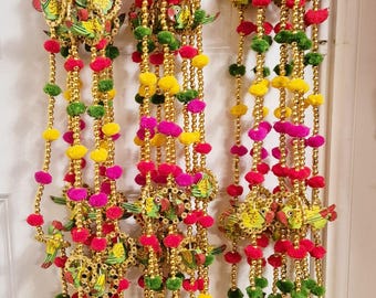 May include: A set of three colorful door hanging decorations with gold bells, pom poms, and small wooden birds. The decorations are made with a variety of colors, including pink, yellow, green, and gold.