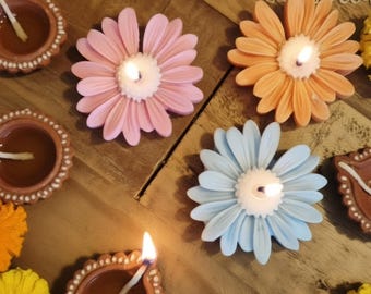 May include: A collection of lit flower-shaped candles in pink, orange, and light blue, interspersed with brown clay oil lamps and vibrant yellow and orange marigold flowers arranged on a wooden surface with a white lace doily.