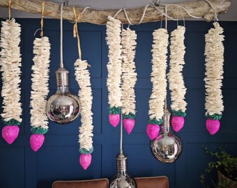 May include: Decorative hanging lights with white fabric strands, pink bulb-shaped accents, and silver light bulbs. The lights are suspended from a natural branch against a dark blue wall. Home decor.
