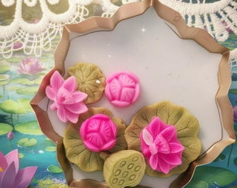 May include: A copper-colored metal cookie cutter shaped like a lotus flower, containing several small, molded soap or wax pieces in the shapes of pink lotus blossoms, green lily pads, and a tan lotus seed pod.