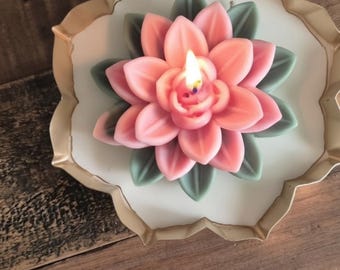 May include: A lit flower-shaped candle with pink petals and green leaves, resting on a white, scalloped-edge plate with a gold-colored rim. The candle is positioned centrally, with the flame burning from the center of the pink petals.