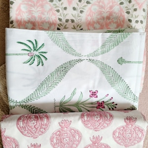 May include: Three fabric swatches featuring different patterns. The top swatch is pink with a white and green floral design. The middle swatch is white with a green palm tree and fern design. The bottom swatch is white with a pink pomegranate design.