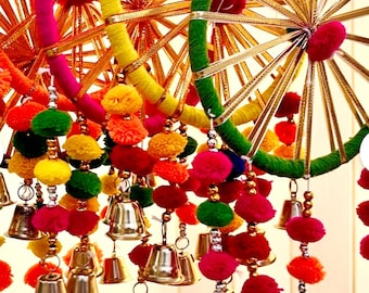 May include: A colorful hanging mobile with multiple circular hoops decorated with yarn and gold trim. The hoops are adorned with pom-poms in various colors, including red, pink, yellow, green, and orange. Bells hang from the bottom of the mobile, adding a whimsical touch.