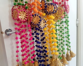 May include: A colorful door hanging with multiple strands of pom-poms in pink, green, yellow, orange, and blue. Each strand has a gold bell at the bottom and a decorative round piece in the middle.