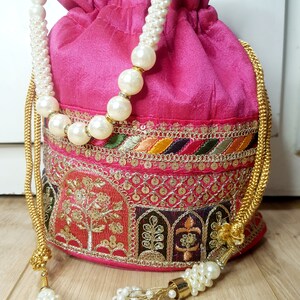 Puede incluir: Una bolsa de cord&oacute;n rosa con bordados dorados y plateados y una cadena de perlas y oro. La bolsa tiene un borla en el extremo de la cadena.