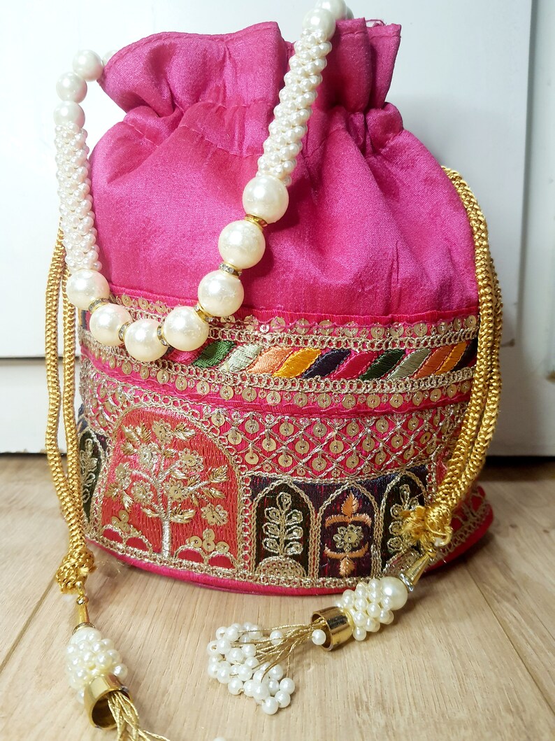 Puede incluir: Una bolsa de cord&oacute;n rosa con bordados dorados y plateados y una cadena de perlas y oro. La bolsa tiene un borla en el extremo de la cadena.