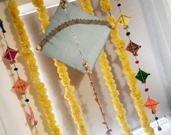 May include: A blue kite with gold trim hangs from a string with yellow marigold garlands and colorful paper houses. The garlands are strung on a metal rod.