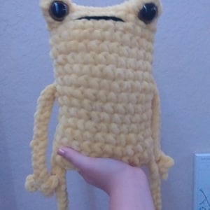 May include: A crocheted yellow frog plush toy with black eyes. The frog has long arms and legs.