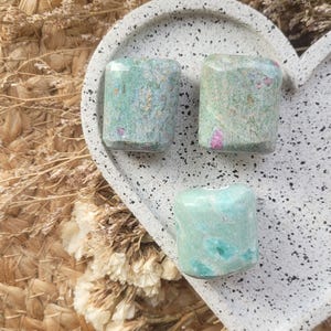 May include: Three green and pink tumbled stones on a white speckled heart-shaped tray. The stones are smooth and have a natural, polished finish.