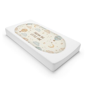 Könnte beinhalten: Eine weiße Wickelauflage mit einem ovalen Mittelteil mit einem skurrilen Design. Das Design umfasst Heißluftballons, Wolken, Sterne und Monde in sanften Pastellfarben. Der Text "DREAM BIG LITTLE ONE" ist in der Mitte aufgedruckt.