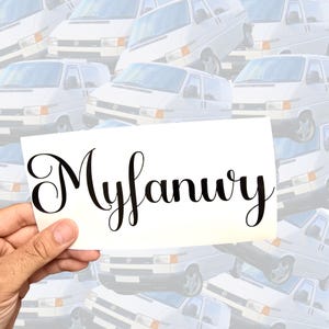 May include: A white rectangular decal featuring the name Myfanwy written in a black, elegant, cursive script font. The lettering is centered on the surface, showcasing a smooth, flowing typeface with distinct, decorative loops on the capital M.
