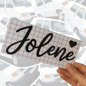 May include: A white sheet of paper with a blue grid pattern. The name "Jolene" is written in black cursive lettering with a small black heart to the right of the name.