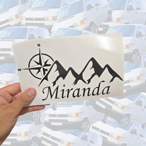 May include: A black vinyl decal on a white backing sheet featuring a compass rose with N and W directional markers, a mountain range silhouette, and the name Miranda written in a cursive script font below the graphic design elements.