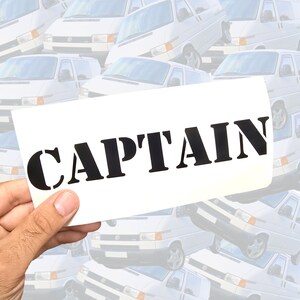 May include: A rectangular white decal featuring the word CAPTAIN printed in bold black capital letters in a stencil-style font. The decal is held against a background pattern of multiple white vans shown from various angles.