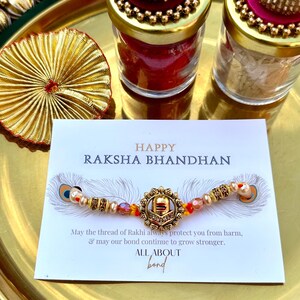 Może przedstawiać: Zestaw prezentowy Raksha Bandhan z ozdobną bransoletką z centralnym wisiorkiem i koralikami, a także małymi słoiczkami z ozdobnymi wieczkami. Zestaw zawiera kartę z napisem "HAPPY RAKSHA BHANDHAN".