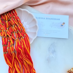 May include: A set of red and orange thread bracelets with gold-toned metallic accents, accompanied by a white card featuring the text HAPPY RAKSHA BHANDHAN and a peacock feather illustration, alongside decorative red and gold circular ornaments.