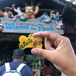 Op de afbeelding: Een gele, botvormige huisdierpenning met een afbeelding van Winnie de Poeh die een honingpot vasthoudt en de naam Winnie in zwarte letters. De penning heeft een klein gaatje aan de bovenkant voor bevestiging en een glanzende afwerking.