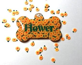 Könnte beinhalten: Eine knochenförmige Haustiermarke aus klarem Harz, gefüllt mit kleinen gelben und orangefarbenen blumenförmigen Konfetti. Das Wort Flower ist in dunkelgrünen Buchstaben in der Mitte aufgedruckt. Lose Blumenkonfetti liegen um die Marke herum.