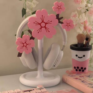 May include: A white headphone stand featuring three pink cherry blossom flower decorations with dark brown stems. The stand has a circular base and a central vertical post that supports a pair of white over-ear headphones.