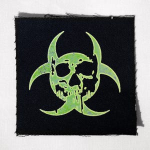 Puede incluir: Parche cuadrado negro con un diseño de calavera verde neón y símbolo de peligro biológico. La calavera tiene detalles de goteo y el parche tiene un borde sin rematar. El diseño está centrado en el parche.