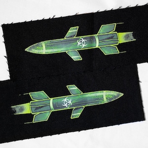 May include: Two black fabric patches with a green and yellow rocket design. The rockets have a biohazard symbol and are detailed with fins and exhaust. The edges of the patches are unfinished.