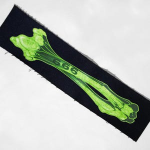 May include: A black rectangular patch featuring a neon green bone graphic with the number "666" printed on it. The patch has a raw edge.