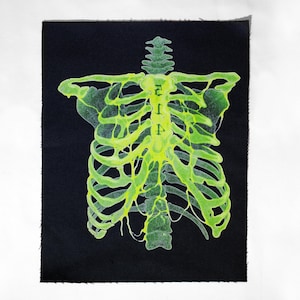 May include: A neon green skeleton design is displayed on black fabric. The rib cage and spine are detailed with a glowing effect. The word "RIP" is vertically printed on the spine. The fabric has a raw edge.