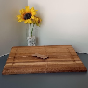 May include: A rectangular wooden cutting board with a grooved edge and a small tag tied with twine. The board is made of light and dark brown wood. A vase of sunflowers sits in the background.