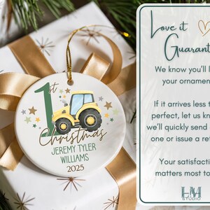 May include: A round Christmas ornament with a yellow tractor illustration, the text "1st Christmas", and the name "Jeremy Tyler Williams" with the year 2025. The ornament is on a wrapped gift with a gold ribbon.