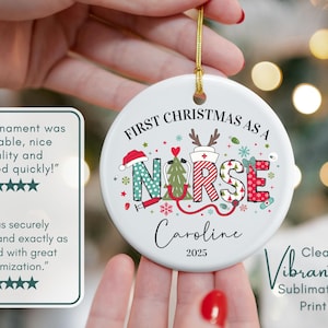 May include: A white ceramic Christmas ornament with the text "First Christmas as a Nurse" and the name "Caroline" and the year "2025". The ornament has a gold string for hanging and features a colorful design with medical symbols.