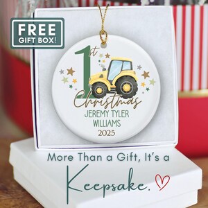 May include: A white ceramic Christmas ornament featuring a yellow tractor with a blue cab, the text "1st Christmas", and personalized details. The ornament is in a white gift box with the text "More Than a Gift, It's a Keepsake."