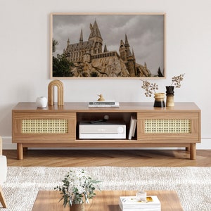 Op de afbeelding: Een lichte houten tv-standaard met twee laden en een plank. De laden hebben een geweven rotan voorzijde. De standaard heeft een donker houten blad en staat op een lichte houten vloer. Een ingelijst afdruk van een kasteel hangt aan de muur boven de standaard.