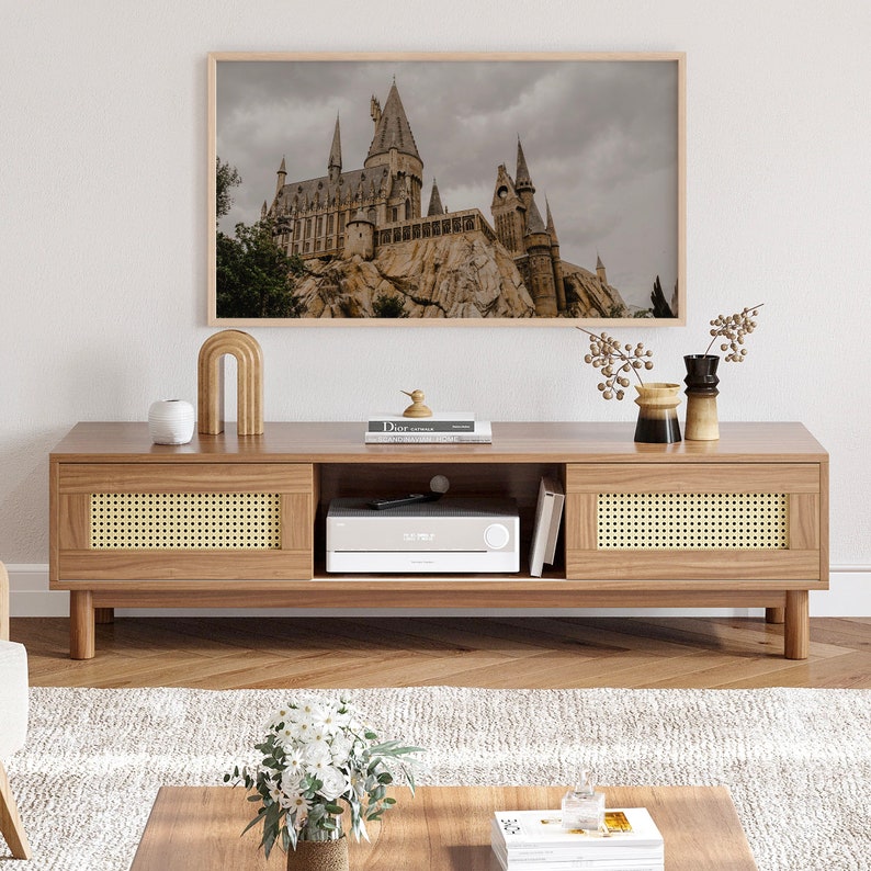 Op de afbeelding: Een lichte houten tv-standaard met twee laden en een plank. De laden hebben een geweven rotan voorzijde. De standaard heeft een donker houten blad en staat op een lichte houten vloer. Een ingelijst afdruk van een kasteel hangt aan de muur boven de standaard.