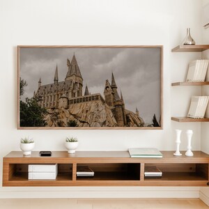 Op de afbeelding: Een ingelijste print van een kasteel met een grijze lucht en groene bomen op de voorgrond. Het kasteel is van steen en heeft veel torens. De print hangt aan een witte muur boven een houten plank met boeken en decoratieve items.