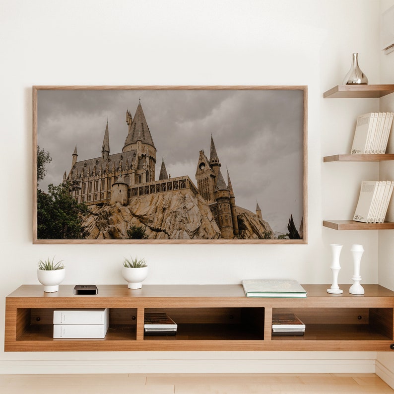 Op de afbeelding: Een ingelijste print van een kasteel met een grijze lucht en groene bomen op de voorgrond. Het kasteel is van steen en heeft veel torens. De print hangt aan een witte muur boven een houten plank met boeken en decoratieve items.