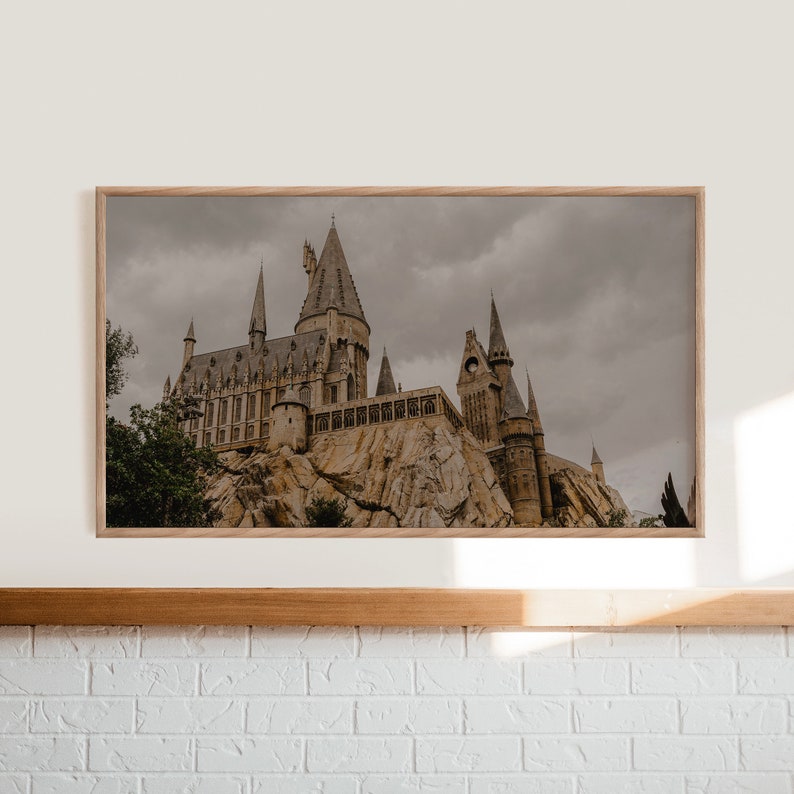Op de afbeelding: Een ingelijste foto van een groot, grijs stenen kasteel met meerdere torens, tegen een bewolkte lucht. Het kasteel staat op een rotsachtige klif, met bomen op de voorgrond.
