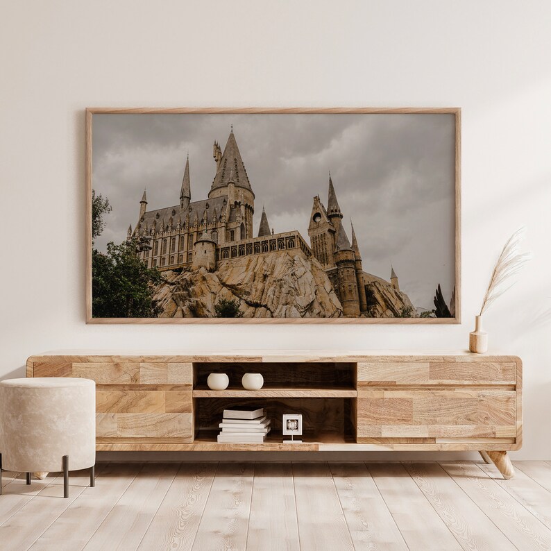 Op de afbeelding: Een ingelijste foto van een groot stenen kasteel met meerdere torens, tegen een bewolkte lucht. Het kasteel staat op een rotsachtige klif, met bomen en groen op de voorgrond. De foto wordt weergegeven op een houten plank met een lichte houtkleur.