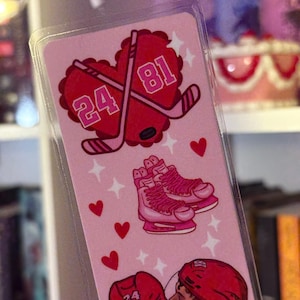 May include: Pink bookmark with hockey-themed illustrations. Features crossed hockey sticks within a heart, numbers 24 and 81, ice skates, and two hockey players kissing. Small red hearts and stars decorate the background.