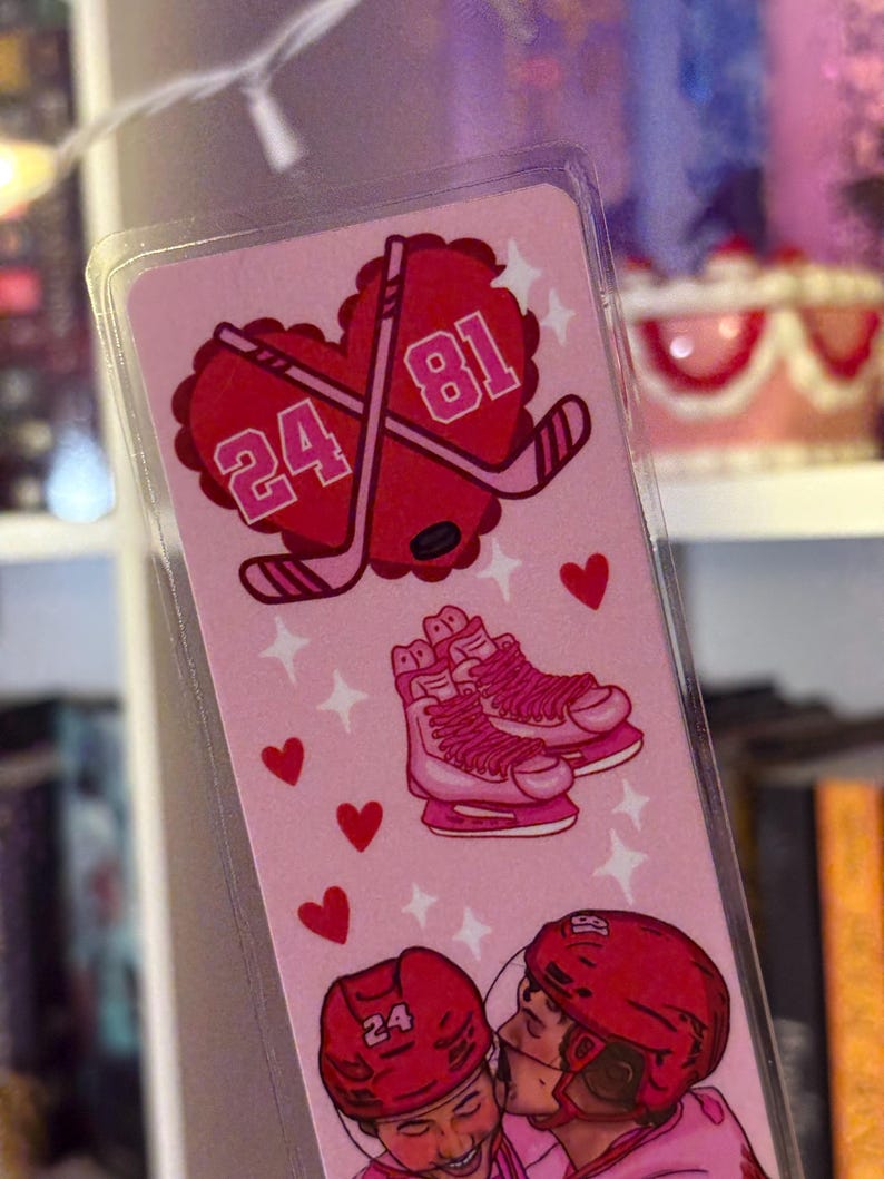 May include: Pink bookmark with hockey-themed illustrations. Features crossed hockey sticks within a heart, numbers 24 and 81, ice skates, and two hockey players kissing. Small red hearts and stars decorate the background.