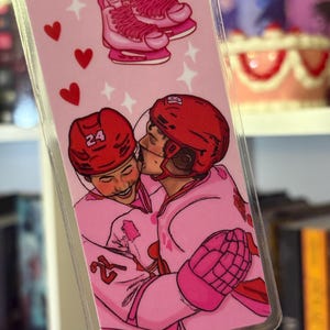 May include: A pink bookmark featuring a hockey theme. The top of the bookmark shows pink ice skates. Below, two hockey players in pink uniforms are embracing. Red hearts and stars decorate the background.