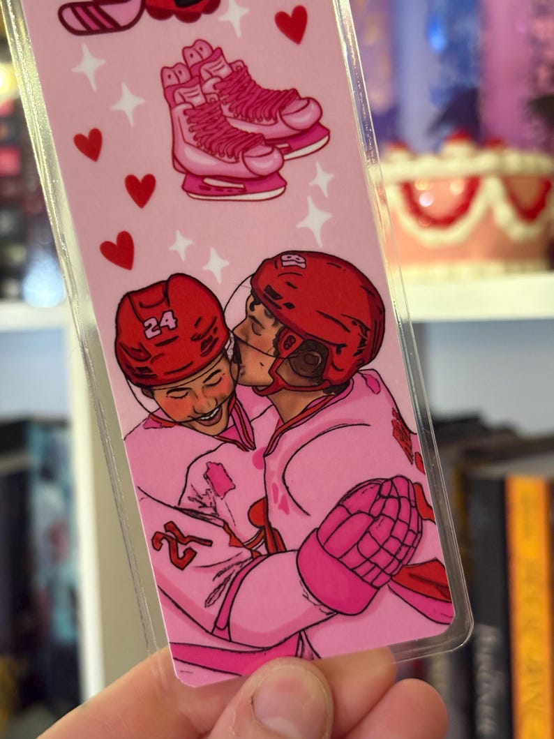 May include: A pink bookmark featuring a hockey theme. The top of the bookmark shows pink ice skates. Below, two hockey players in pink uniforms are embracing. Red hearts and stars decorate the background.