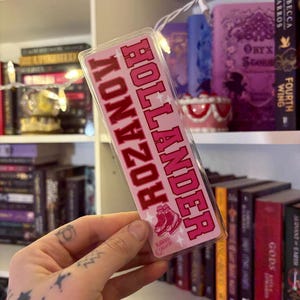 May include: A pink and red bookmark with the text "ROZ HOLLANDER" in a collegiate font. The bookmark features an illustration of pink roller skates and stars. The background includes a bookshelf with various books.