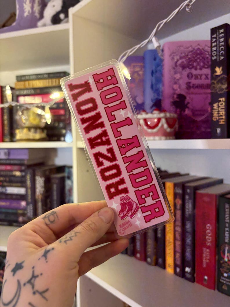 May include: A pink and red bookmark with the text "ROZ HOLLANDER" in a collegiate font. The bookmark features an illustration of pink roller skates and stars. The background includes a bookshelf with various books.