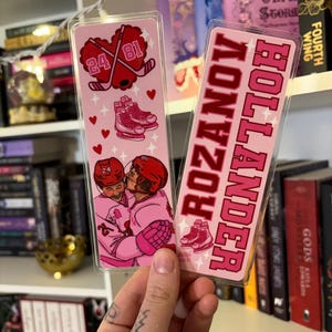 May include: Two pink hockey-themed bookmarks. One features a heart with crossed hockey sticks and the numbers 24 and 81. The other bookmark has the text "Rozanov Hollander" in pink. Both have illustrations of hockey skates and players.