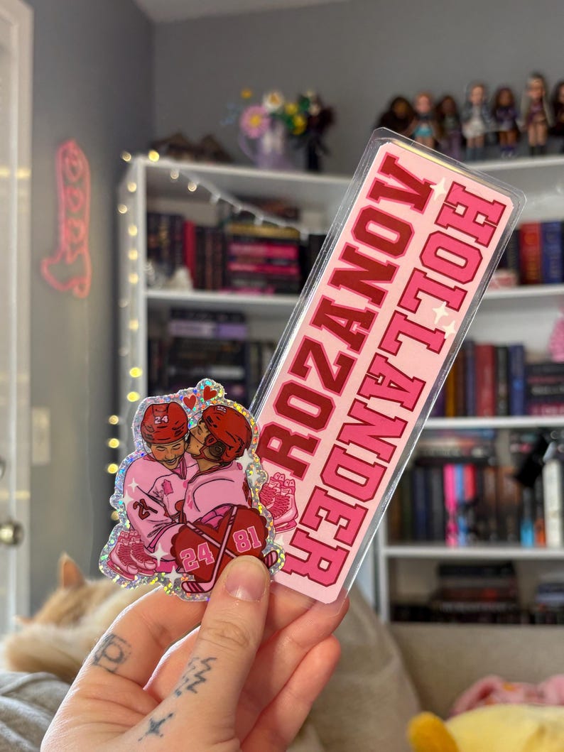May include: A pink sticker with the words "ROZANOV HOLLANDER" in red, and a smaller sticker featuring two hockey players in pink uniforms kissing. The smaller sticker has the numbers 24 and 81 on it.