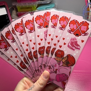 May include: A stack of pink hockey-themed cards featuring a heart-shaped design with crossed hockey sticks, the number 24, and a pair of skates. The cards also depict two hockey players embracing. Small red hearts and stars decorate the cards.